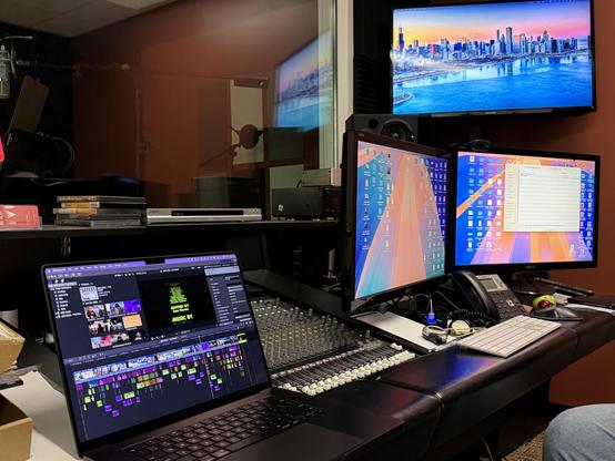 A recording studio with a mixing console, computer screens, and a microphone. A cityscape is displayed on a TV screen.