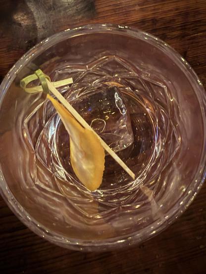 A clear glass with a large ice cube and a yellow peel on a wooden surface.