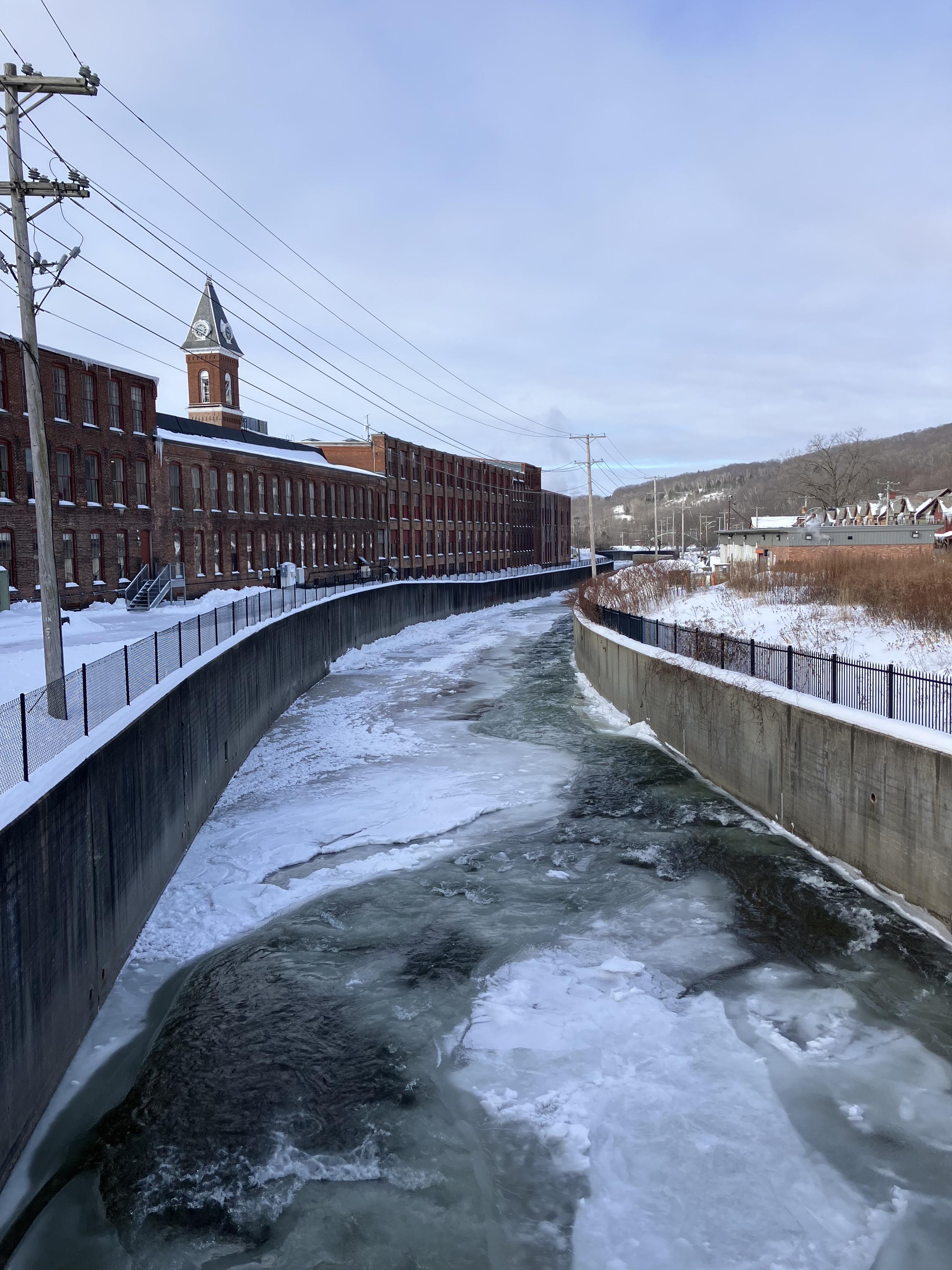 Photo of water channel partly frozen over with brick building to the left.