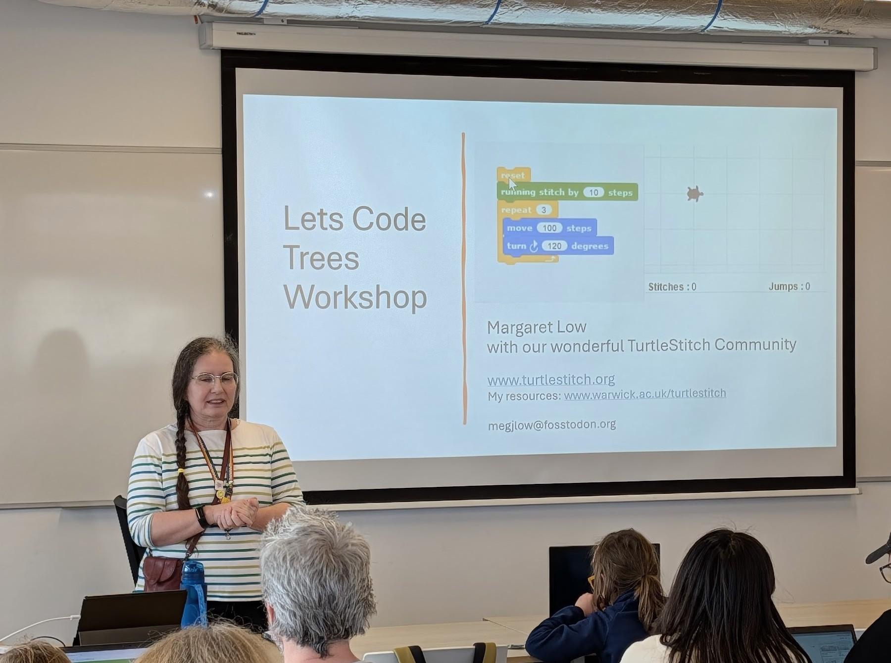 Margaret standing in at the front of a classroom, beside a display screen, showing the title "Lets Code Trees Workshop"