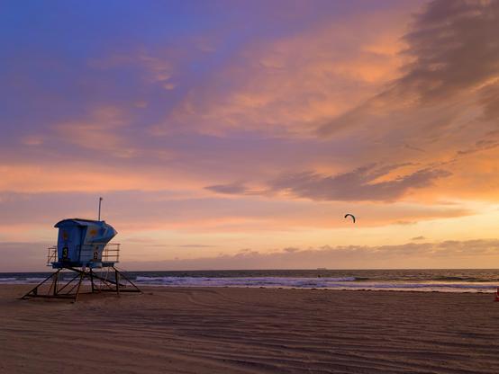 A blue lifeguard tower on a sandy beach under a colorful sunset sky with a person parasailing in the distance.