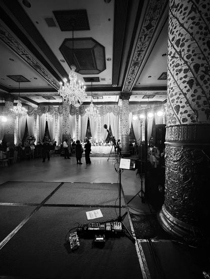 A black and white photo of a ballroom with chandeliers, columns, and a stage with audio equipment.