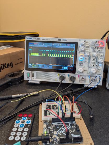 A black Metro Arduino next to a white solderless breadboard with a circuit containing wires, push buttons, resistors, LED's and a transistor. Behind this is an oscilloscope displaying somewhat square digital signals in yellow and blue. A mini remote control is next to the Metro.
