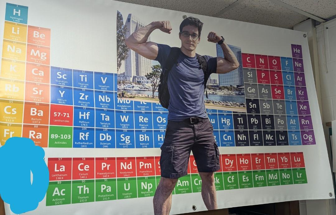 A photo of me in front of a periodic table printed on a large vinyl banner. My arms have been replaced by a much more muscular set as a joke. 