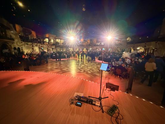 A dance floor with people dancing under colorful lights. A stage with a microphone and sound equipment is in the foreground.