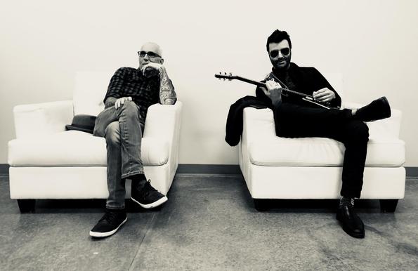 Two men in sunglasses sit in armchairs. One plays a guitar, the other rests his chin on his hand. Both wear casual, dark clothing.