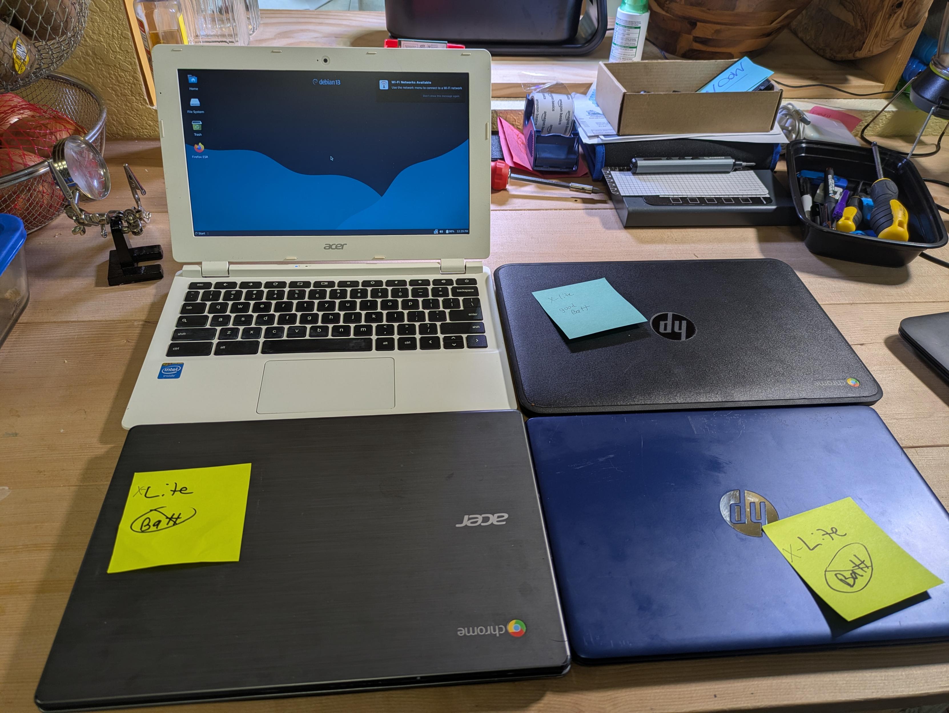 4 older Chromebooks sitting in a desk. One is open showing a Debian 13 xfce desktop 