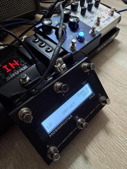 A black pedalboard with a digital display and several knobs and switches. There are cables connected to the pedals.