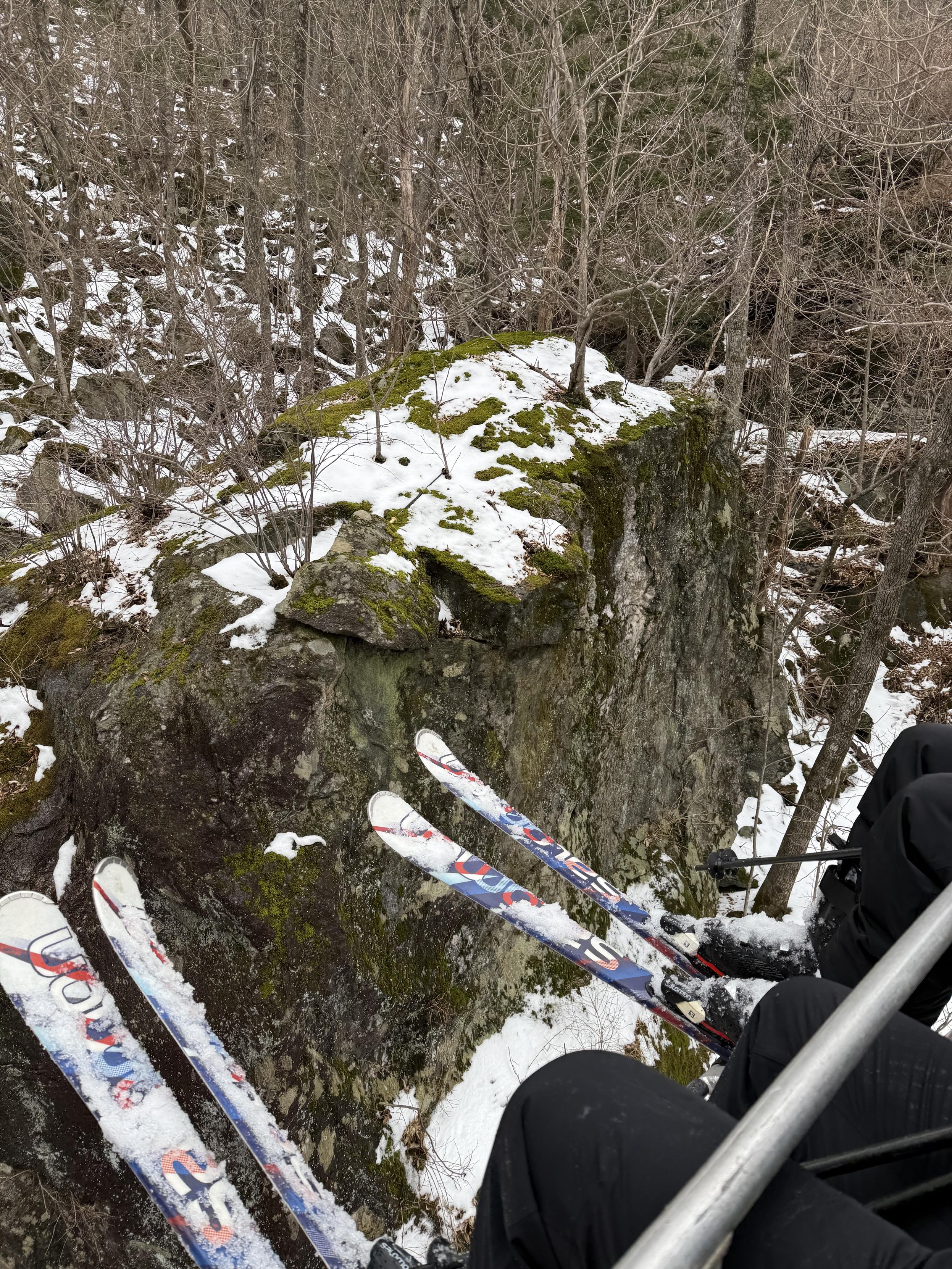 Several riders in a ski lift passing close to a blunder with light snow and moss cover. Several small trees can be seen growing on top.