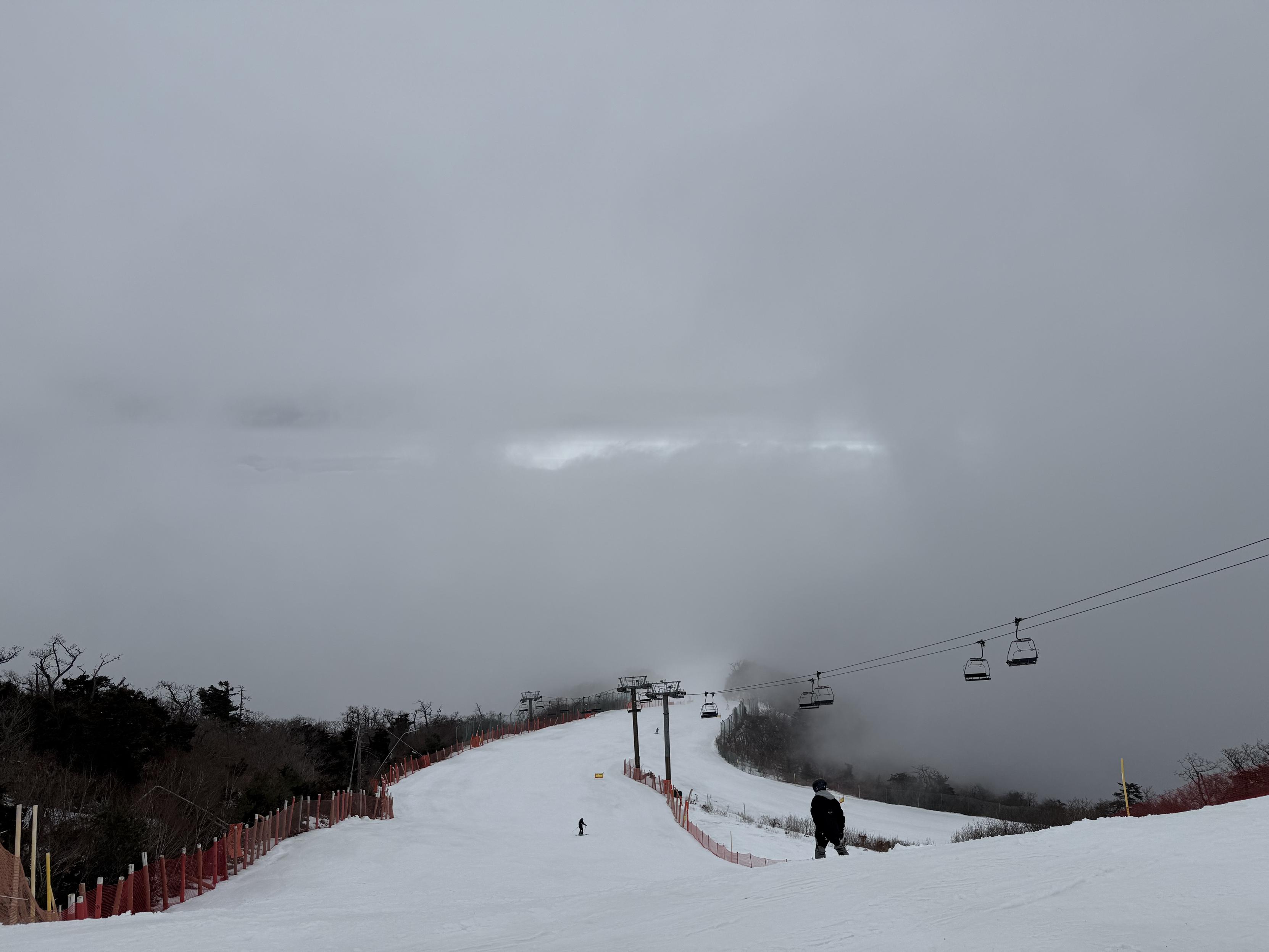 View from the top of a ski slope. You can’t see more than a hundred or so meters into the distance due to fog. The sky has clouds of varying thickness. A patch of light is peeking through one of the less dense sections.