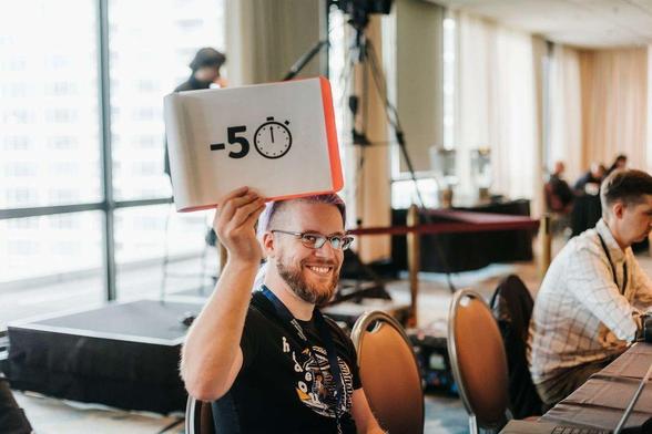 Session chair Eric Sherman holds up a sign reading -5 with a stopwatch icon at DjangoCon US 2025