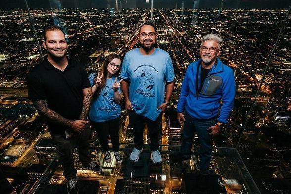 DjangoCon US 2025 attendees with the city of Chicago's night lights behind them as they stand in a high glass observation deck.