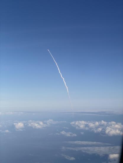 View out of the window of an aeroplane of the trail of smoke from the Artemis rocket against the blue sky, far above the clouds