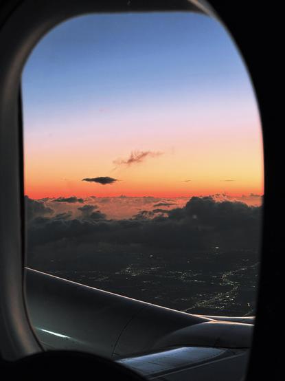 View out of an airplane window at dawn. The sky fades from dark blue to orange, bathing the tops of the clouds with a warm glow. Below the clouds, it's still night, and on the ground we see the lights of the towns.