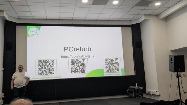 Jon standing in front of a large screen, on the screen is the url for pcrefurb organisation. https://pcrefurb.org.uk
