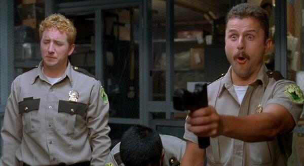 Screenshot from the film Super Troopers, with Mac holding his service weapon up with a mischievous look on his face.