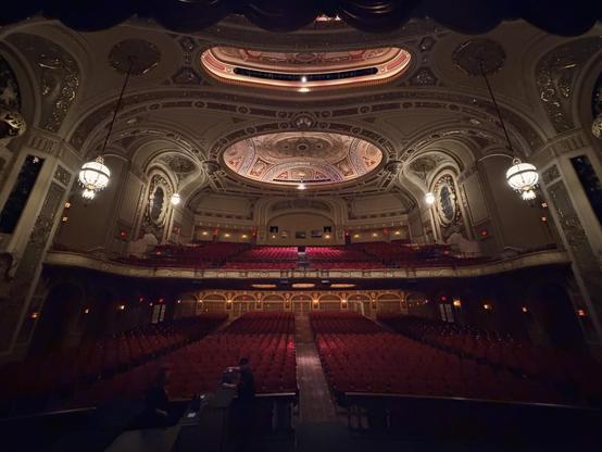 Looking out from the stage onto the beautifully decorated theatre space, with red velvet chairs on the main floor and in the balcony, and beautiful chandeliers hanging down.