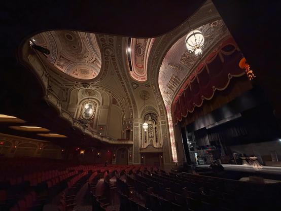View from one side of the theatre space, with the stage on the right and the balcony in the upper left. We can see all the beautiful detailing of the ceiling, as well as the red velvet curtain, and seats.