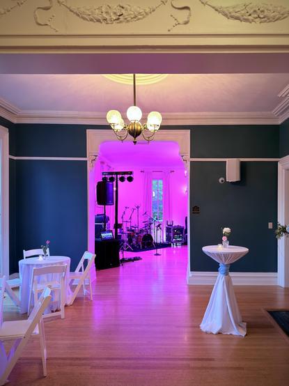 View through a drawing room of an old Victorian mansion, hardwood floors, white tables and chairs set up on the left, a white cocktail table on the right, Pacific blue walls, with beautiful crown mouldings and a six globe pendant in the centre. Through an archway, On the other side of the room, illuminated in pink light, we see the band set up