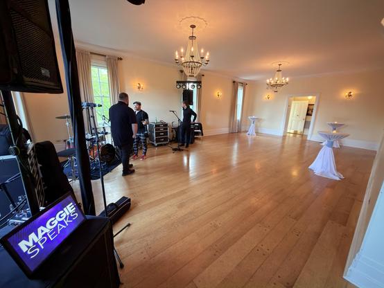 Wide angle view of a larger space in a Victorian mansion, hardwood floors throughout, a few cocktail tables with cinched white draperies on them, band equipment visible on the left, with a small screen displaying the Maggie Speaks logo, chandeliers hanging from the ceiling