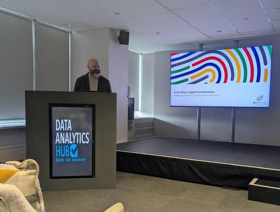 Bearded guy standing behind a podium that says "Data Analytics Hub", with a screen showing a slide entitled South Africa's Digital Transformation 