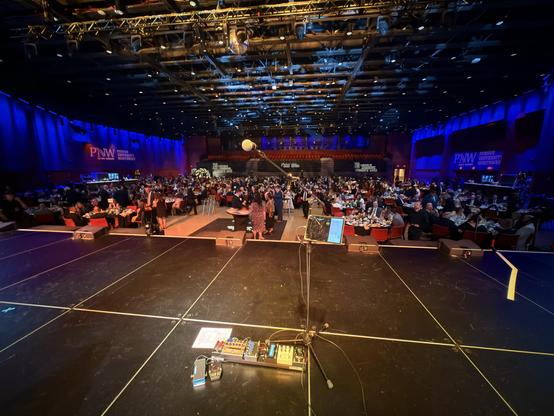 View from the stage at the Hard Rock Casino in Gary IN. My pedalboard and mic stand in the centre of a pretty huge stage, with the large space in front lit by blue and orange lights. We see several hundred guests seated at large round tables, with black tablecloth and red chairs.