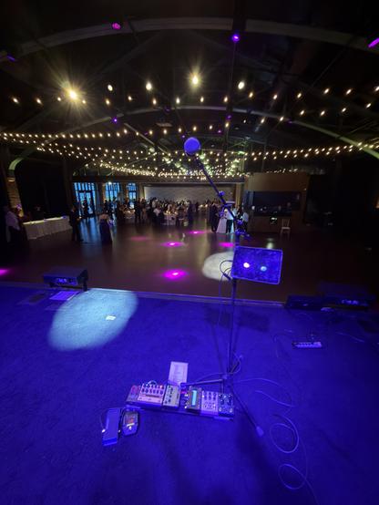 My pedalboard and microphone stand on stage, bathed in blue light, with a pair of white spotlights sweeping the floor. Looking out on an event space with string lights hung, and a small gathering of wedding guests at the other end of the room.