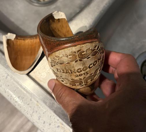 A close-up photograph shows a hand holding a large, broken piece of a ceramic mug over a metallic sink. The mug piece features a dark brown rim and a cream-colored section decorated with dark brown swirling patterns. Centered on the cream-colored section, the text "DJANGO CON 2015" is printed. A second broken fragment of the mug is visible to the left.