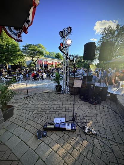 My Mike stand and paddleboard on a brick paver area, small PA and lighting rig in front of me, edge of a pavilion visible on the top left, clad in red white and blue bunting. In front of the stage area there are folding chairs and tables set up in front of a big red barn, with audience milling around everywhere