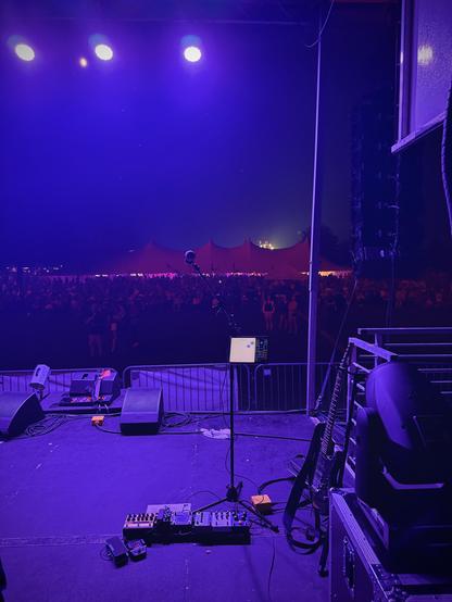 My pedal board on a festival stage, bathed in purple light. Beyond the edge of the stage we see metal barricades, then the crowd lit dimly in the red glow of fireworks. A festival tent looms in the background.
