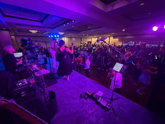 Wide angle view of the band performing from my side of the stage in a banquet hall, under blue and purple lights. The dancing crowd is waving lighted foam sticks around
