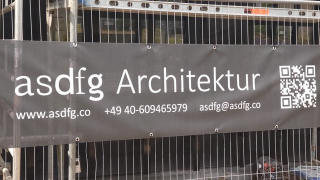 A black banner on a construction fence, reading

asdfg Architektur
www.asdfg.co

...