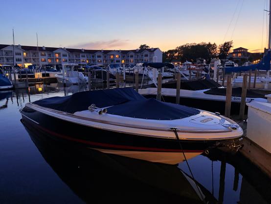 A classic wooden speedboat sits on mirrorlike water in a marina at dusk