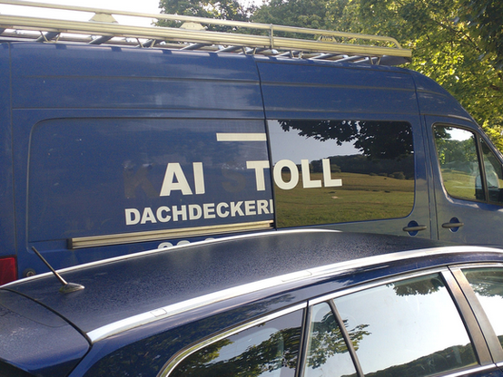 blaues Handwerkerauto, auf der Seite ist in weißen Buchstaben zu lesen "AI TOLL DACHDECKER" (eigentlich KAI STOLL DACHDECKERMEISTER, aber einige Buchstaben sind offenbar schwarz oder ausgeblichen und somit kaum sichtbar)
