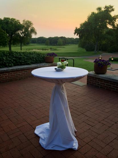 A round high top table, dressed in white, with a couple of small flowers in the centre, sits on a red brick patio, overlooking the greens of a golf course at sunset.