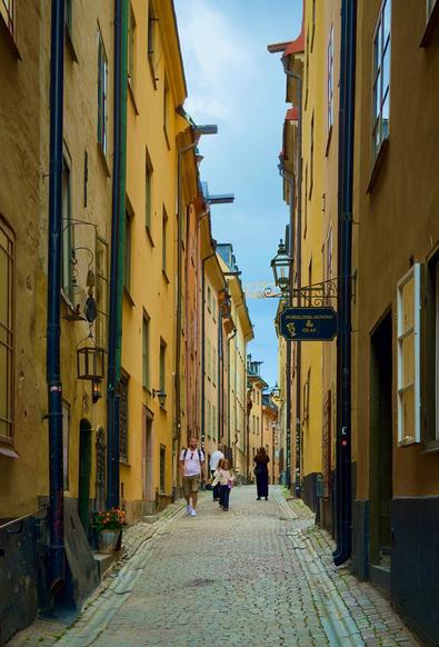 A father and child amble along a narrow medieval cobblestone street, lined with buildings  painted various shades of yellow
