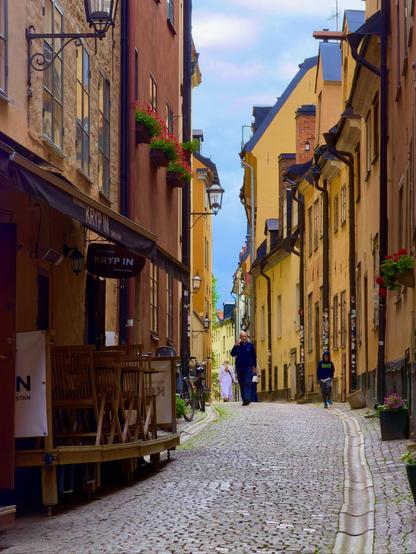 Passersby amble along a narrow cobbled street, lined with buildings in various shades of yellow, a wooden cafe awning visible in the foreground.