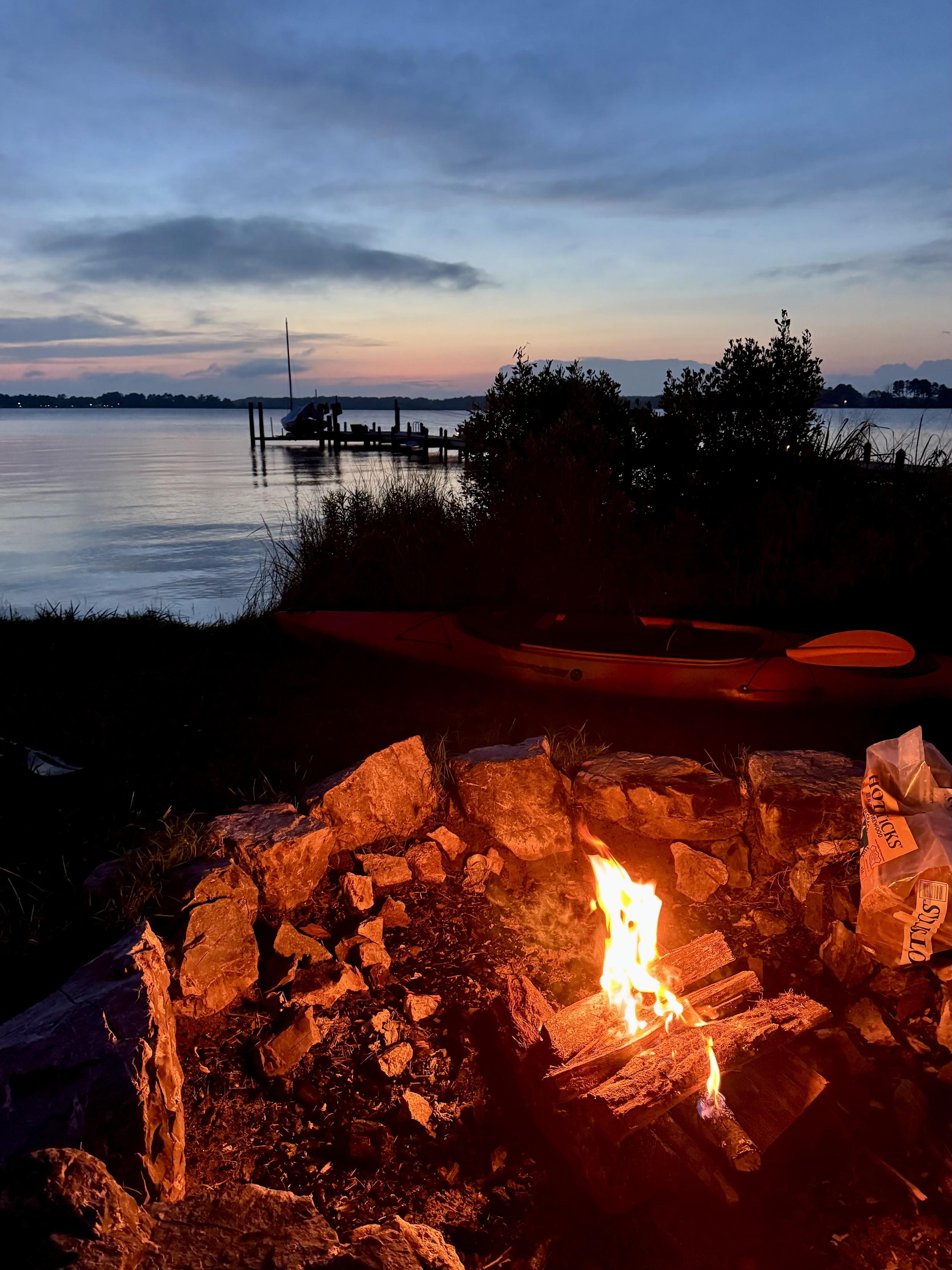 Small campfire in a stone ring on the shore, sunset and pier reflecting in calm water.