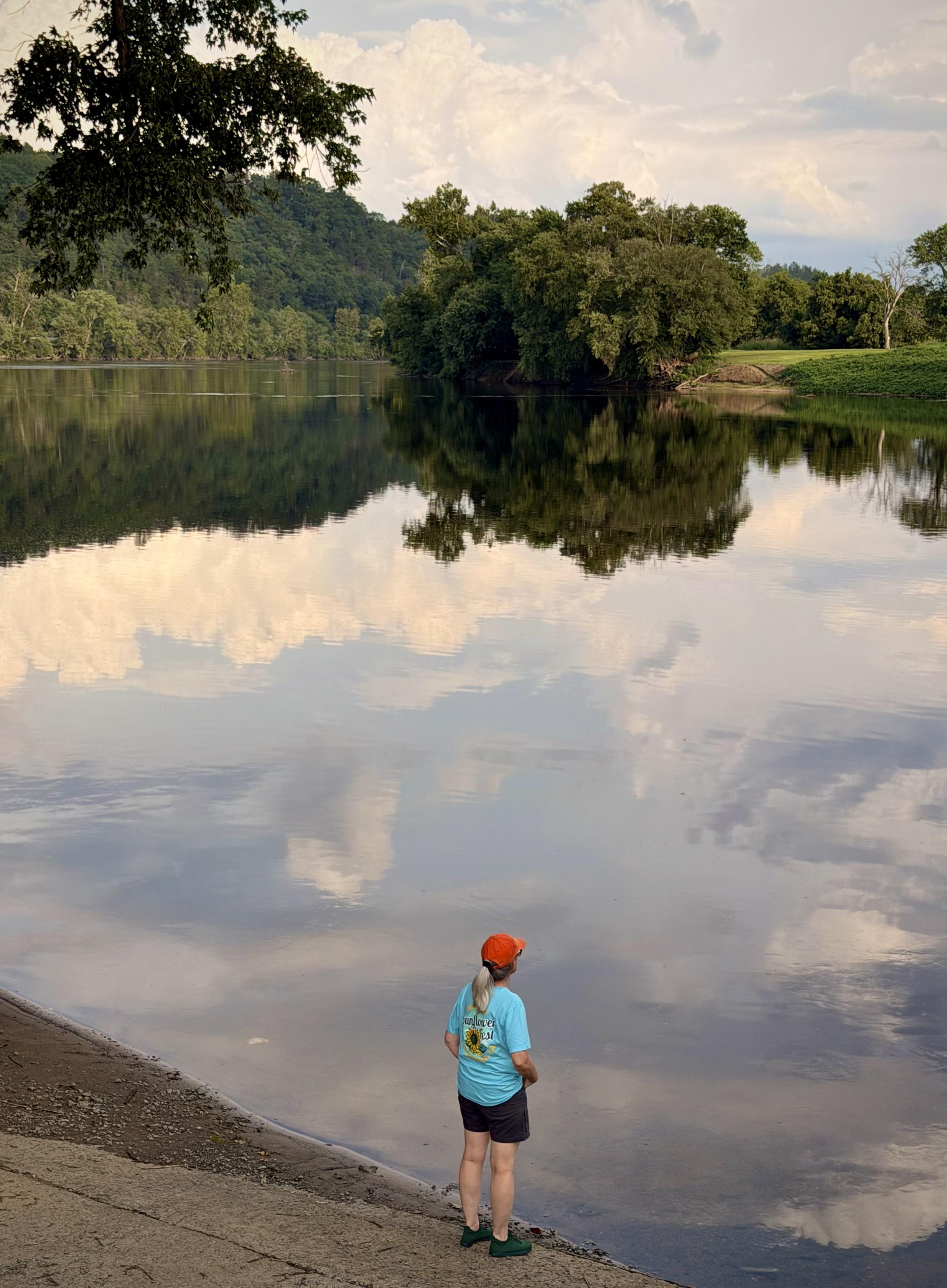 A woman stands at waters edge reflecting evening sky and clouds in wide calm river.