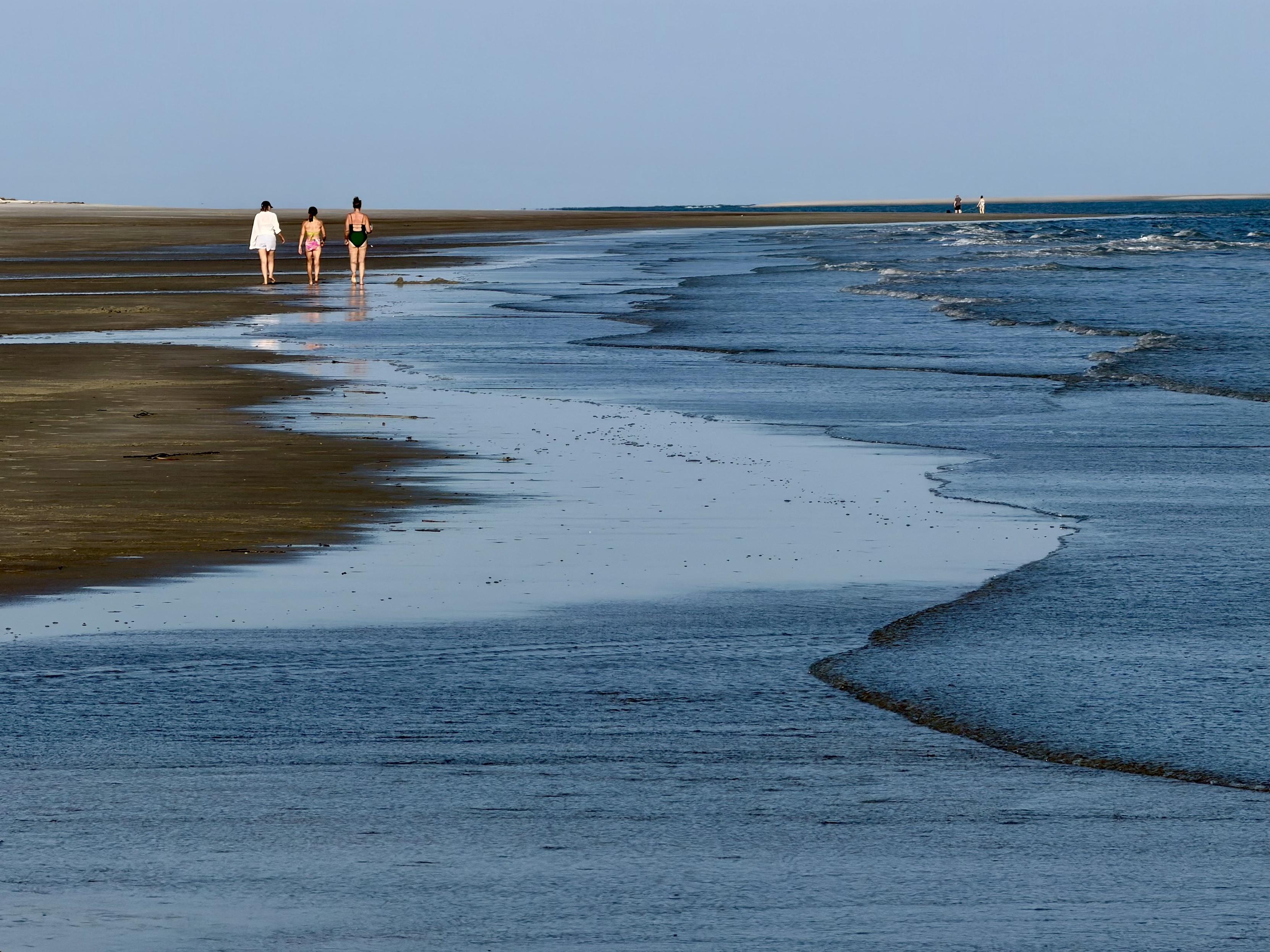 Three women walk in the distance on a wide flat shore along the water’s edge