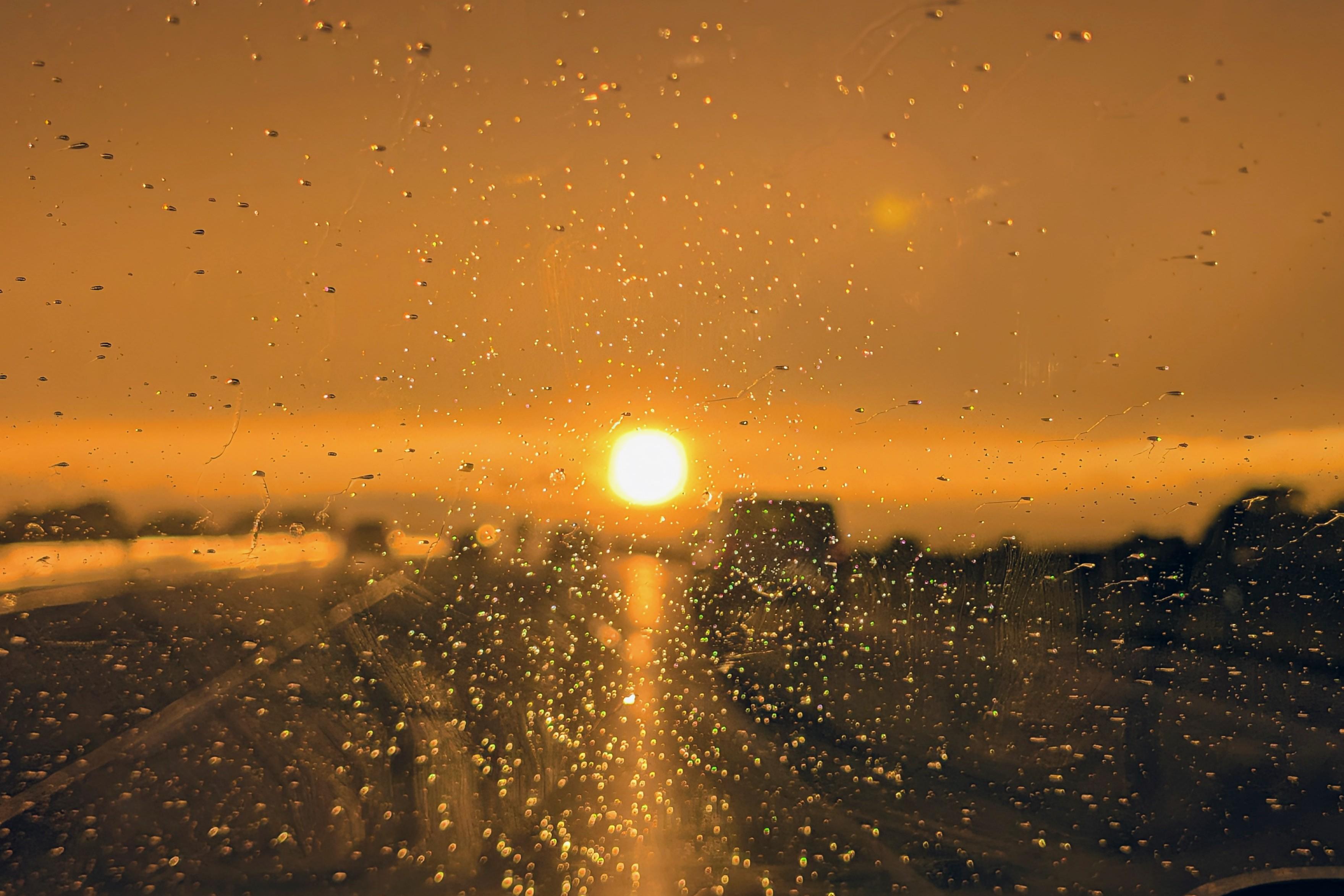 Sunset seen through a rain-covered windshield, warm golden-orange light, water droplets in the foreground.