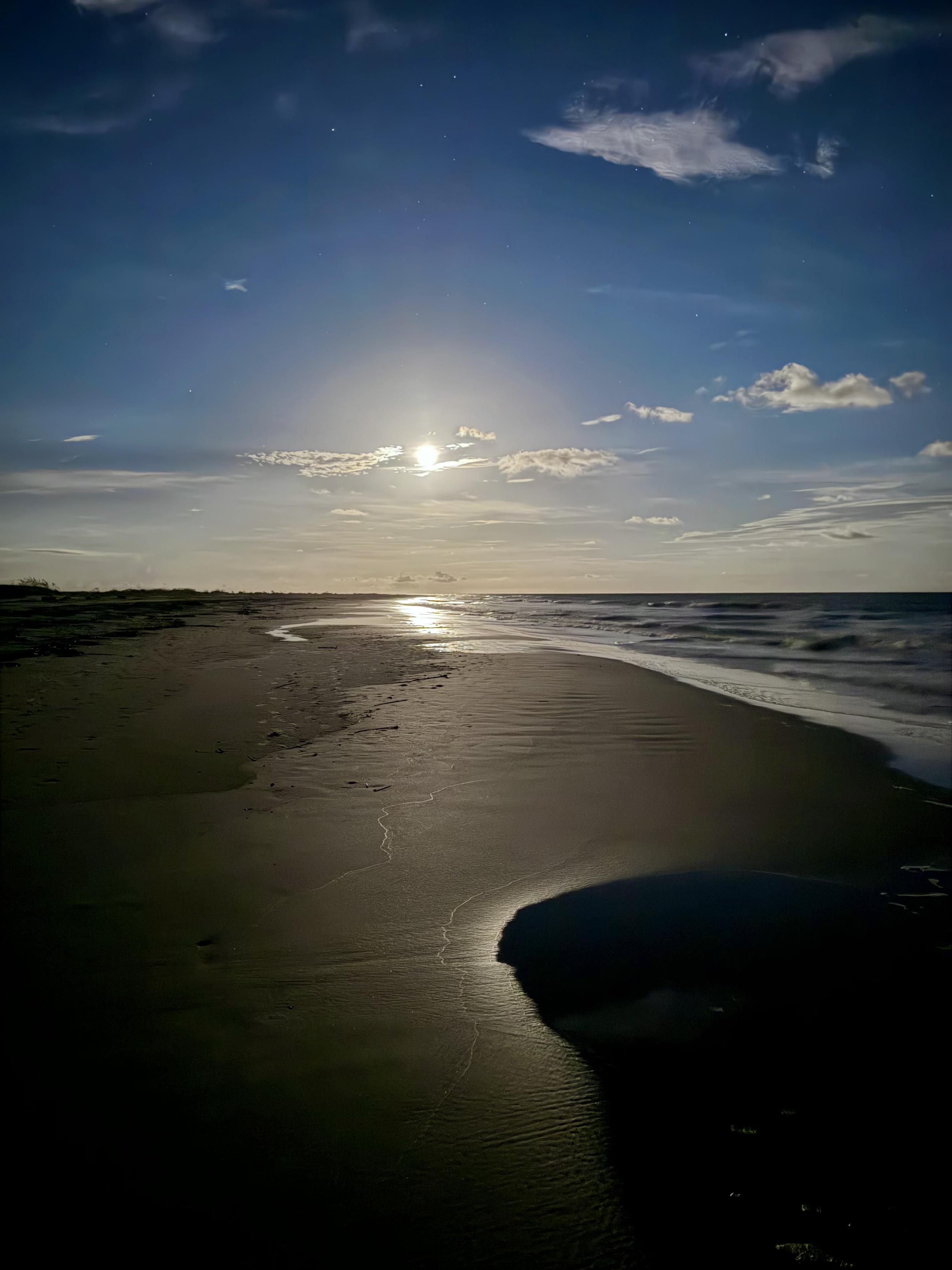 Full moon rising over a beach with flat sand and surf.