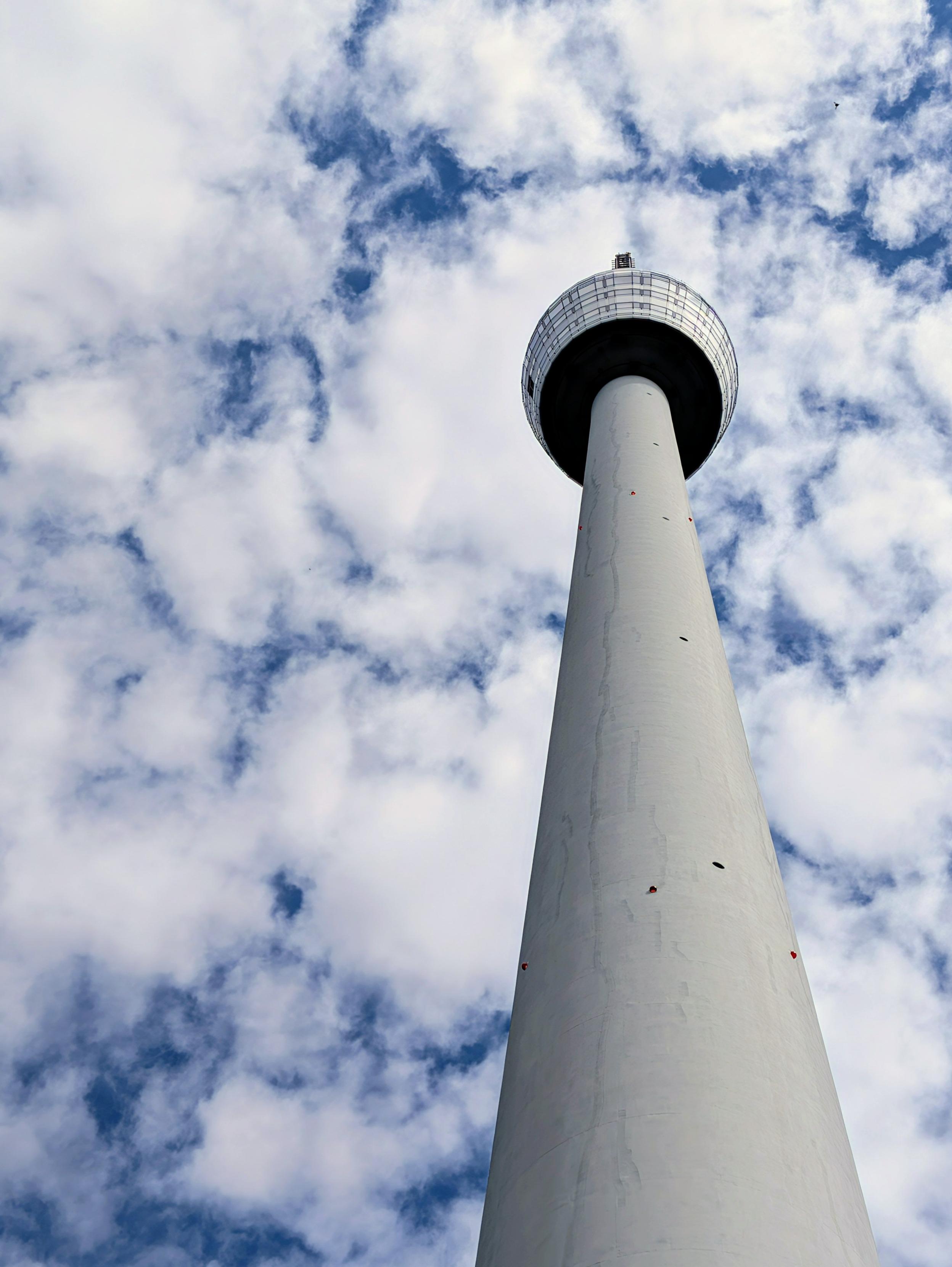 Looking straight up at the Stuttgart TV Tower: the slim white concrete shaft rises steeply into a blue sky dotted with small white puffy clouds. At the top sits the distinctive tower basket with its observation deck.