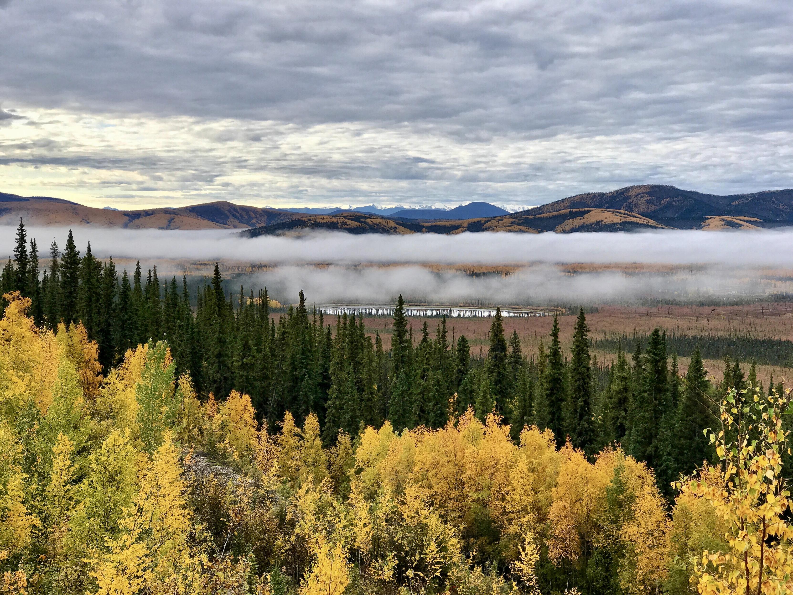 View across a fig filled valley with fall foliage and evergreens and a lake reflecting the sky, mountains in the distance.