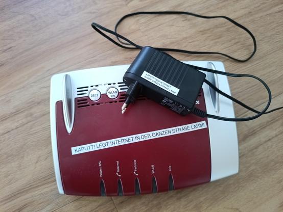 unplugged FritzBox with power supply on top, both sticker-labeled in German with "BROKEN! WRECKS THE ENTIRE STREET'S INTERNET"