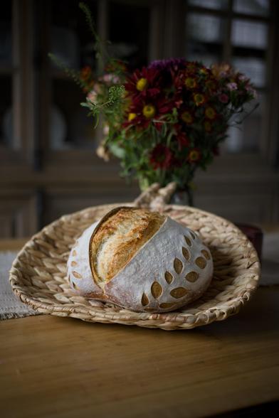A beautiful loaf of bread sits on a pumpkin-shaped basket bathed in the light from the window. The basket is leaned up against a bouquet of fall flowers, sitting on a wooden table.