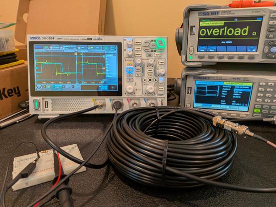 An electronics bench with a coil of coaxial cable and a small breadboard with a 47-ohm resistor. Test leads go to an oscilloscope displaying a pair of square(ish) waves.
