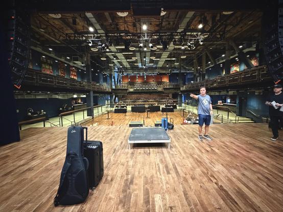 A man points on a wooden stage with a guitar case and suitcase, an empty venue with balconies and overhead lights.