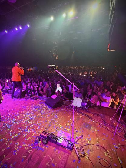 A performer in red sings on a confetti-strewn stage under purple lights, facing a large crowd.
