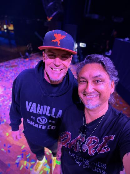 Two smiling men pose on a confetti-covered floor under purple light; one wears a red-winged cap and hoodie.